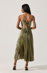 A woman wearing a shimmering, metallic gold pleated dress with thin spaghetti straps. The dress features an open back with a tie detail at the waist, creating cut-out sections on either side. She is also wearing black high-heeled shoes with ankle straps.